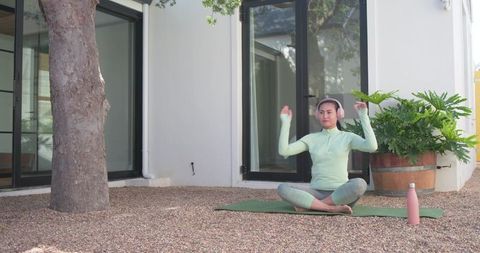 Mid adult asian woman practicing outdoor yoga on patio adjusting pink headphones