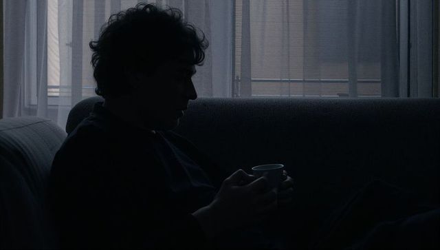 Silhouetted man sitting on sofa holding mug by curtained window in moody blue-gray light