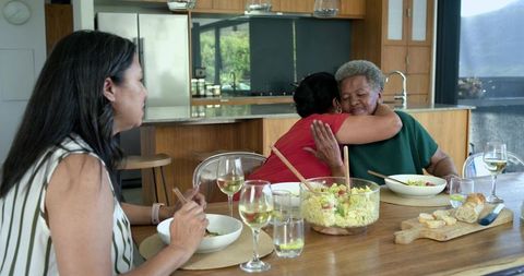 African american multigenerational women hugging at kitchen table sharing salad and wine
