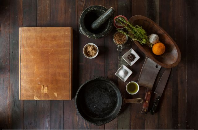 Overhead view of rustic kitchen setup features chopping board, cast iron pan, herbs, spices, garlic, pepper, and fresh orange. Ideal for use in culinary blogs, cookbooks, gourmet recipe illustrations, and food preparation demonstrations. Emphasizes autentic, natural cooking environment.