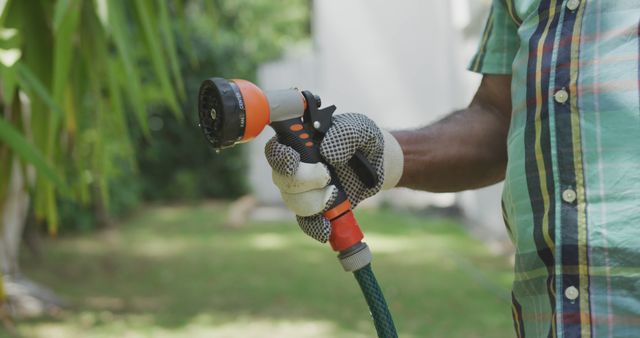 A gardener wearing checkered shirt and gloves holding garden hose with spray nozzle. Suitable for gardening, home improvement, or lawn care themes. Perfect for articles on gardening tips, summer yard maintenance, and eco-friendly watering techniques.