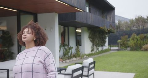 Woman standing on modern patio wearing pink grid sweater looking toward lawn and garden