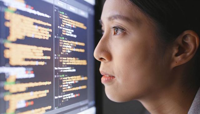 Asian woman studying code on large monitor, focused developer working in dim workspace