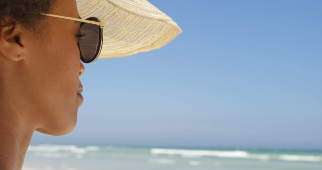 A young African American woman wearing sunglasses and a sunhat is gazing out at the sea, with copy space. Her profile against the beach backdrop evokes a sense of summer relaxation and tranquility.
