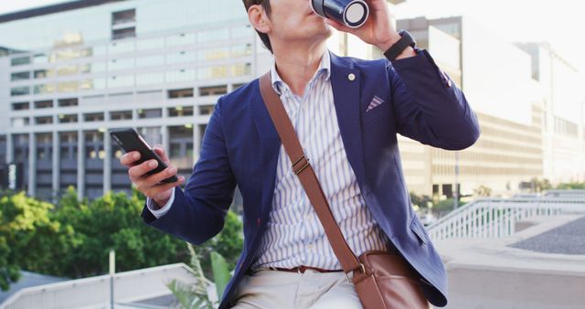 Businessman drinking coffee and using smartphone outdoors with a modern city building in the background. Ideal for depicting modern business lifestyle, professional work environment, remote working, and urban professionals. Suitable for projects related to corporate life, business communications, and outdoor workspaces.