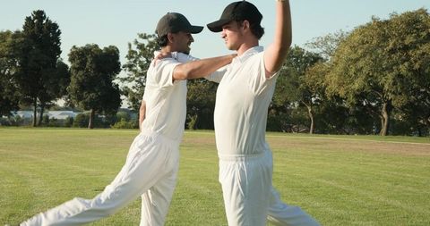 Two cricketers stretching and balancing on grass field during team warmup session