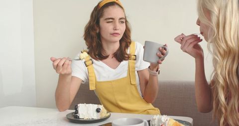 Young women sharing cake and coffee in cozy dining nook, playful wink and casual moment