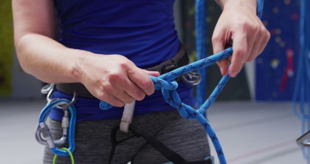 A person tying a knot in a climbing rope while wearing a harness at an indoor climbing gym. Useful for illustrating topics related to rock climbing, safety in sports, proper techniques, climbing equipment, and indoor recreational activities.
