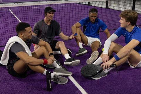 Multicultural men sitting on purple padel court resting after match holding paddles