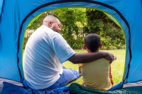 African american father hugging son inside blue tent, camping together in green park