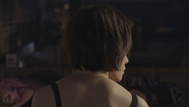 Young woman adjusting black strap top, bob haircut, moody bedroom, intimate evening