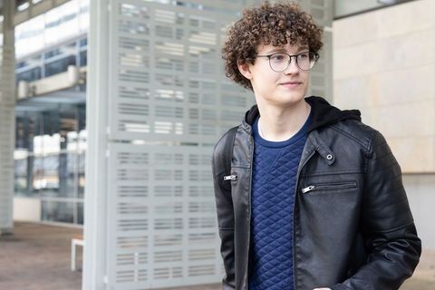 Curly-haired teenage student standing near modern plaza wearing leather jacket and backpack