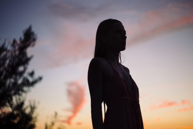 Silhouette of woman standing outdoors against vibrant sunset sky with trees in background. Highlighting serene, peaceful moment in natural setting. Perfect for campaigns about relaxation, contemplation, nature, or introspection. Suitable for blogs, websites, posters, and emotional storytelling visual content.