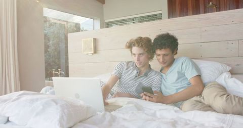 Diverse Gay Couple Relaxing on Bed with Laptop and Smartphone