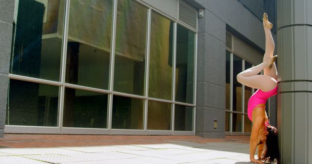 Young female athlete is training by performing a handstand against a column in an urban setting. She is wearing a bright pink athletic outfit and white leggings. The background features modern building architecture with glass panels. This can be used for themes related to fitness, athleticism, urban workouts, flexibility exercises, and promoting active lifestyles.