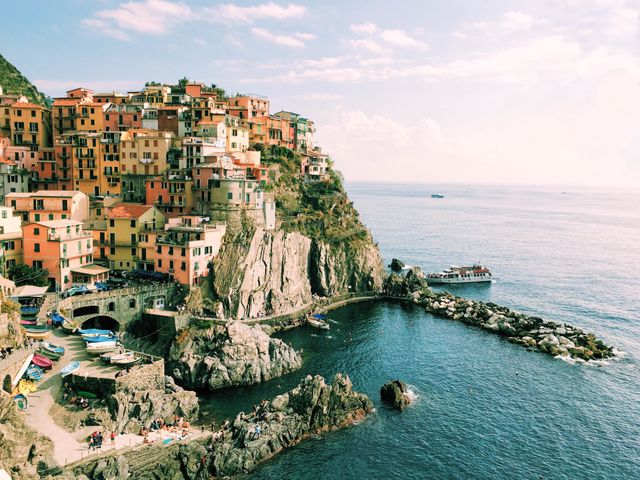 Colorful buildings sit atop a rocky cliff overlooking the sea in a picturesque coastal village, likely in Italy's Cinque Terre. The scene features a sunny day, with a boat near the waterfront and clear blue skies. Ideal for promoting tourism, travel destinations, scenic illustrations, and Mediterranean lifestyle concepts.