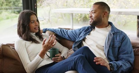 Multiethnic couple having cozy conversation on leather couch with coffee and natural light