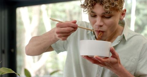 Young Man Enjoying a Bowl of Noodles with Chopsticks Near Window