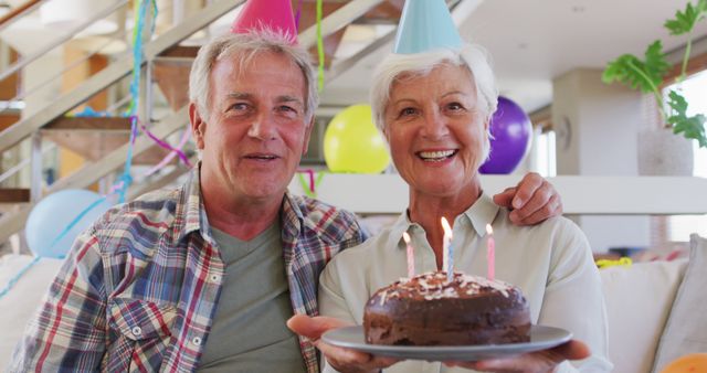 Senior couple celebrating a birthday together at home with a chocolate cake and candles. They are wearing party hats and smiling happily, surrounded by balloons and festive decorations. Ideal for use in promotional materials for birthday celebrations, senior living communities, and family events.