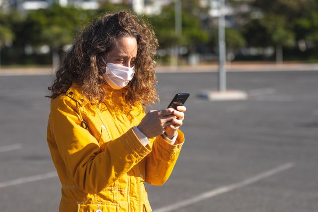 Caucasian woman standing on pavement in city during daytime, wearing face mask for protection against COVID-19, using smartphone. Perfect for illustrating pandemic safety measures, urban lifestyle, technology use, and health precautions.