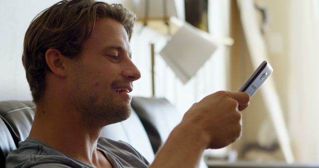 Young man using his smartphone while sitting on a couch indoors. He is smiling, suggesting he is enjoying a text conversation or content online. Ideal for use in projects about communication, technology, everyday life, or casual indoor activities.