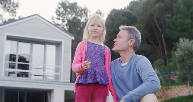 Image of a father and his young daughter spending quality time outdoors in front of a modern house. The daughter is smiling confidently, standing near her father who is sitting, gazing at her affectionately. The house has a contemporary design with large windows. This image can be used for themes related to family life, real estate, parenting, and suburban living.
