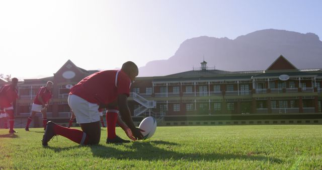 Rugby players in red jerseys on a sunlit field preparing for a game with a school building and mountains visible in the background. Ideal for promoting team sports, athletic training, school rugby programs, or teamwork concepts.