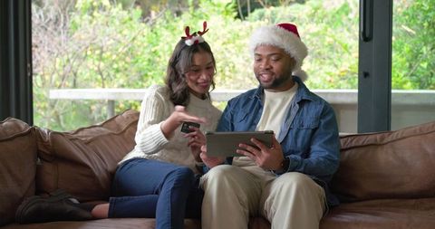 Multicultural couple celebrating christmas shopping on tablet from cozy leather couch