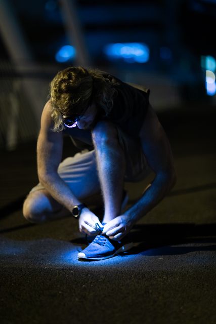 Front view of a fit Caucasian man with long blonde hair wearing sportswear exercising outdoors in the city at the evening, kneeling and tying his shoe, wearing head light.