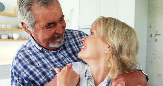 A senior Caucasian couple shares a tender moment, with the man embracing the woman lovingly in a home setting. Their affectionate gaze reflects a deep connection and years of companionship.