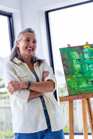Senior woman artist standing and smiling beside easel displaying green abstract canvas