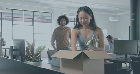 Young woman packing desk lamp into moving box in modern open-plan coworking office