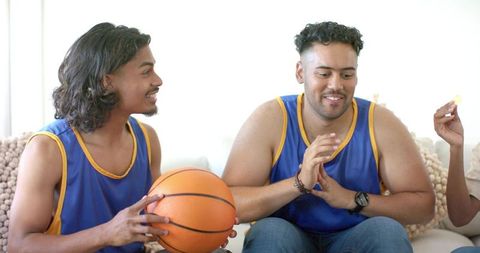 Friends cheering on sofa in matching blue basketball jerseys sharing chips and ball