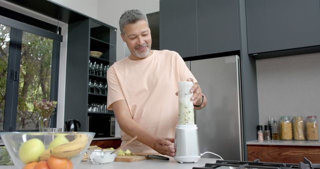 Middle-aged man preparing ingredients and using blender in modern kitchen. Perfect for themes on cooking, healthy living, food preparation, or kitchen appliances.