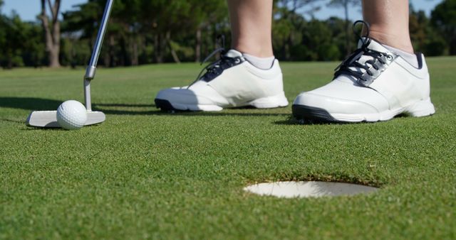 Focus on golfer's white shoes and putter while putting golf ball close to the hole on greens of golf course. Perfect for advertisements, sports articles, golf tutorials, and promotional materials for golfing events and equipment.