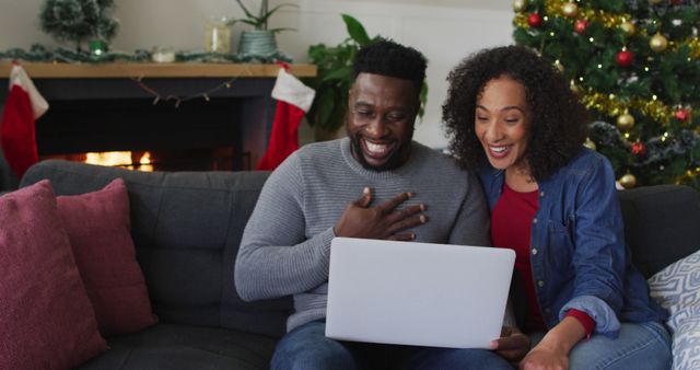 Couple sitting on sofa using laptop for a video call, enjoying Christmas. Fireplace and decorated Christmas tree in background. Uses include holiday greeting cards, content on virtual holiday celebrations, family bonding themes, and seasonal marketing.