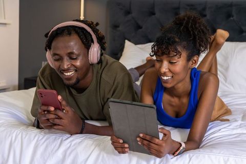 African american couple relaxing on bed using tablet and smartphone, wearing headphones