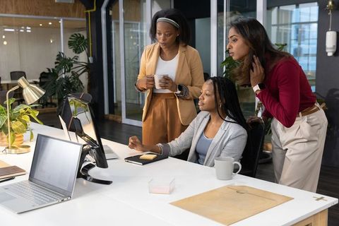 Diverse women collaborating at modern office desk, black and south asian team brainstorming