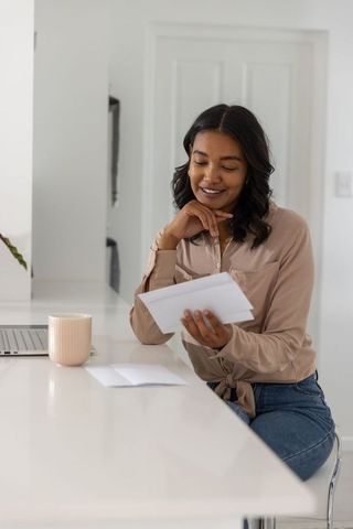 African american woman reading letter at kitchen counter with laptop and mug