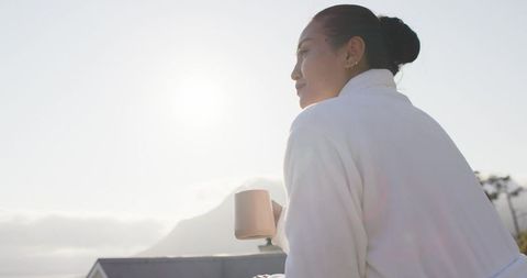 Asian woman enjoying sunrise on rooftop balcony wearing white robe, holding mug, gazing out