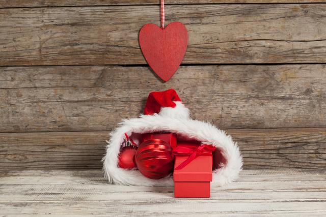 Christmas baubles and a gift are placed inside a Santa hat with a heart decoration hanging above. The wooden background adds a rustic feel, making it perfect for holiday-themed promotions, greeting cards, festive advertisements, and seasonal blog posts.