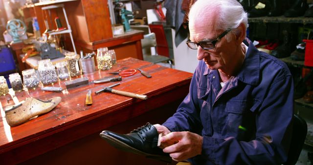 Elderly cobbler is repairing shoes at his wooden workbench. Workspace includes various tools, jars of nails and a shoe last. Ideal for portraying traditional craftsmanship, manual labor, or artisanal trades.