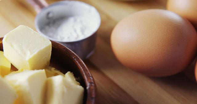 Close-up view focused on baking ingredients on a wooden tabletop. Butter cubes in a bowl, whole eggs, and a measuring cup with flour are displayed. Ideal for content related to cooking, baking blogs, recipe demonstrations, or food preparation tips.