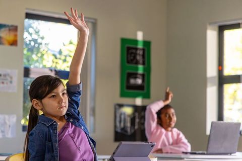Asian girl actively raising hand in classroom with tablet and laptop for hybrid learning