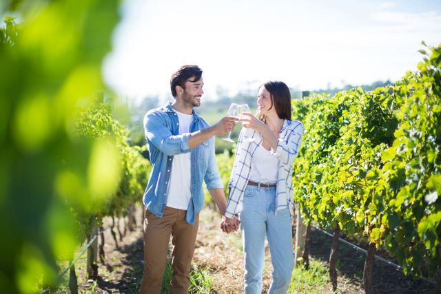 Couple standing in lush vineyard, holding hands and toasting wineglasses. Ideal for use in advertisements for wine brands, romantic getaways, lifestyle blogs, and travel brochures. Perfect for illustrating themes of romance, celebration, and outdoor leisure activities.