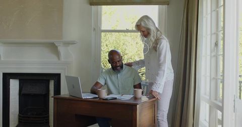 Mature couple reviewing finances at home desk with laptop, paperwork and coffee mugs by window