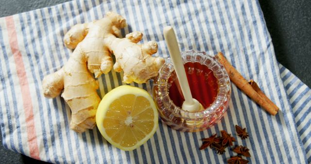 Fresh ginger root, a sliced lemon, and a jar of honey with a dipper are arranged on a striped cloth, suggesting natural remedies or ingredients for a healthy recipe. These items are often associated with wellness and homemade treatments for colds or flu.