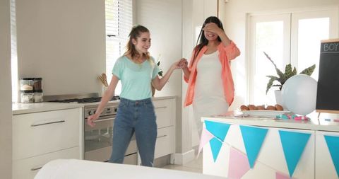 Pregnant woman receiving surprise baby shower reveal guided by friend in sunlit kitchen