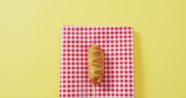 Corn dog topped with mustard placed on red and white checkered napkin, set against yellow background. Ideal for food blogs, culinary websites, picnic-themed promotions, fast food advertisements and cafe menus.