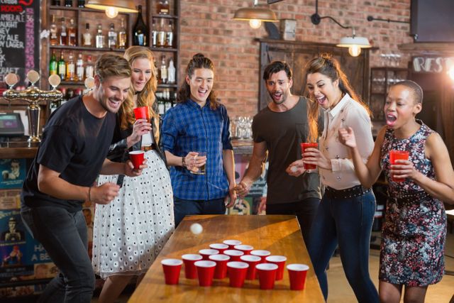 Friends cheering while man playing beer pong on table in bar